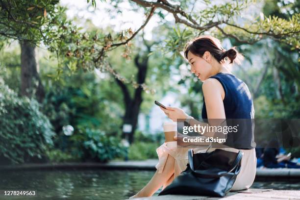 young asian businesswoman taking a coffee break in urban park in the city. she is sitting by the pond, using smartphone and enjoying a cup of coffee in the middle of a work day - woman smartphone nature stockfoto's en -beelden