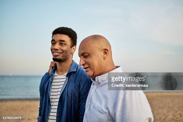 affectionate father walking on beach with young adult son - dad advice stock pictures, royalty-free photos & images