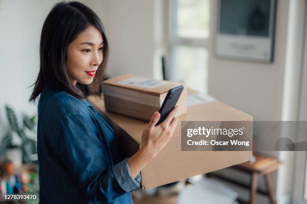 young woman reading message on mobile phone whilst carrying a stack of delivery boxes at home - telephone box stock-fotos und bilder