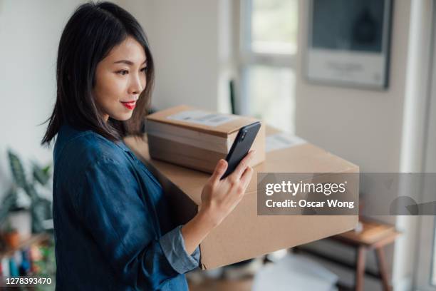 young woman reading message on mobile phone whilst carrying a stack of delivery boxes at home - consegna a domicilio foto e immagini stock
