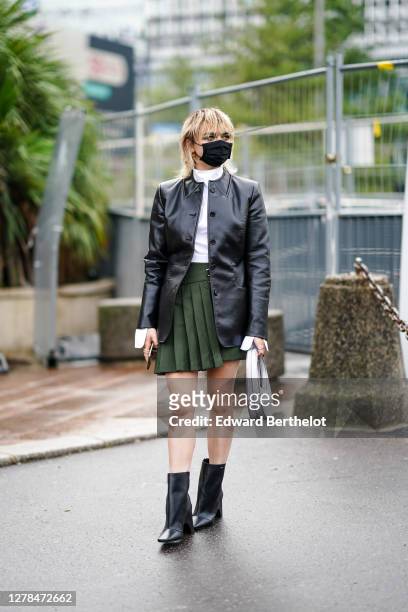 Maisie Williams wears a black protective face mask, a white top, a black leather jacket, a green pleated skirt, black leather pointy boots, outside...