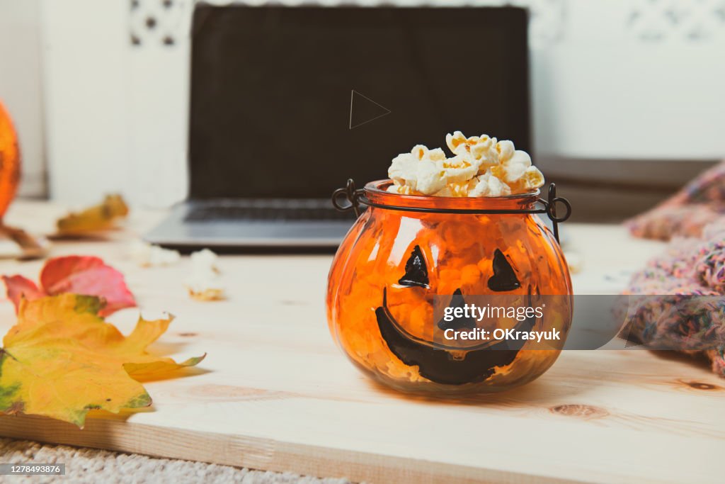 Halloween Pumpkin bowl filled with popcorn with an open laptop, warm plaid and fallen leaves on the wooden background. Halloween Movie party concept. Cozy and safe holiday. Copy space.
