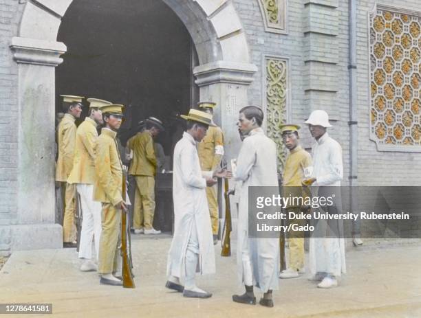 Young men wearing white tunics , and uniformed guards with rifles, stand outside the arched entrance to the Students' Jail, Beijing, China, 1919....