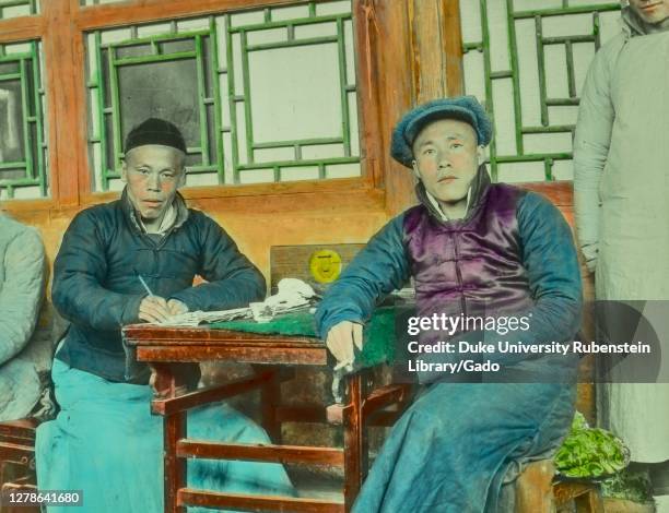 Mid-shot of two businessmen, facing the camera with serious expressions, sitting outside flanking a table covered with papers and a small wooden...