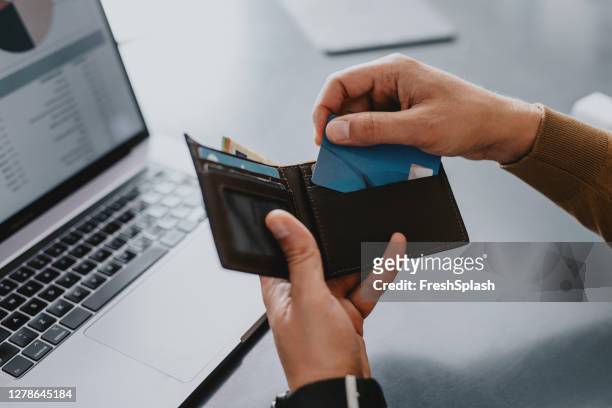 hände eines mannes, der seine kreditkarte aus seinem portemonnaie nimmt, um auf online-bestellung auf seinem laptop-computer zu machen - wallet stock-fotos und bilder