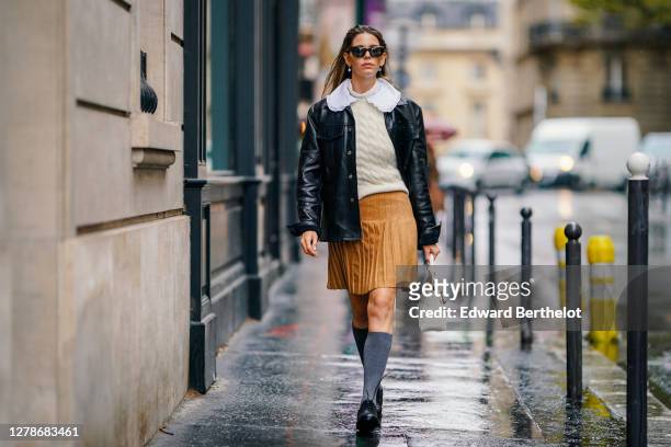 Natalia Verza wears sunglasses, a white wool woven pullover, a white ruffled collar, a brown/orange pleated skirt, a black leather jacket, gray high...