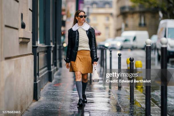 Natalia Verza wears sunglasses, a white wool woven pullover, a white ruffled collar, a brown/orange pleated skirt, a black leather jacket, gray high...