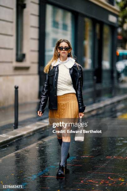 Natalia Verza wears sunglasses, a white wool woven pullover, a white ruffled collar, a brown/orange pleated skirt, a black leather jacket, gray high...