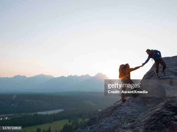 due alpinisti offrono aiuto su una cresta rocciosa all'alba sopra una valle - vetta foto e immagini stock