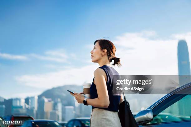 confidence and professional young asian businesswoman walking out of her car, holding smartphone and a cup of coffee. looking towards the sky against urban cityscape. business on the go concept - china oost azië stockfoto's en -beelden