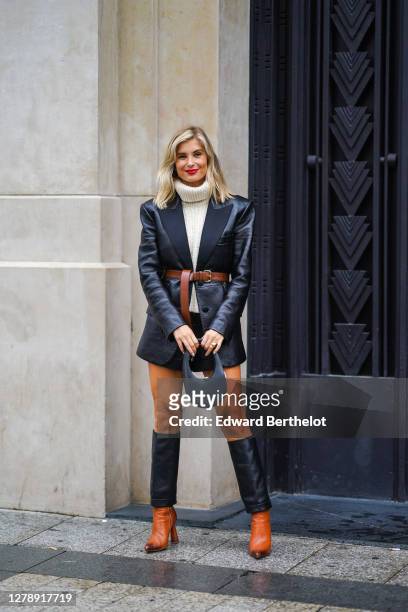 Xenia Adonts wears a white wool turtleneck pullover, a black leather blazer jacket, a brown leather belt, leather shorts, a bag, brown and black...