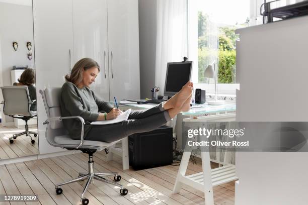 mature woman working at desk at home taking notes - businesswoman barefoot stock-fotos und bilder