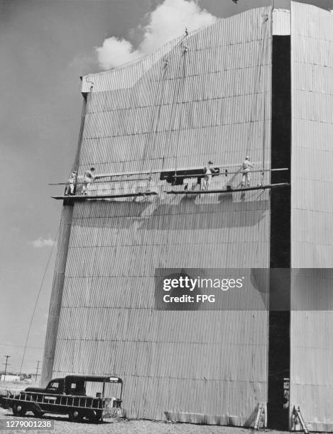 Painters standing on a suspended cradle, painting the words 'truck tires' on the entrance to a warehouse building or hangar, location unidentified,...
