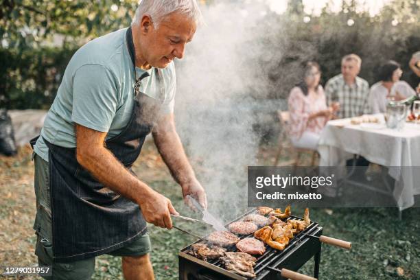 senior man cooking meat on barbecue grill in open garden - family barbeque garden stock pictures, royalty-free photos & images