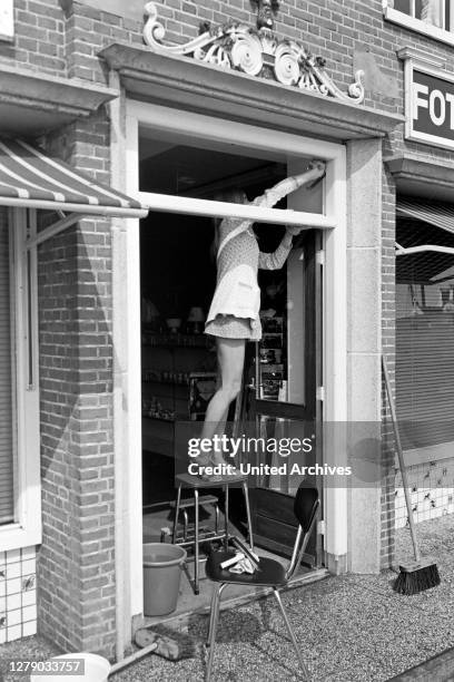 Young woman wearing a mini dress cleaning the windows of a photographic shop at Volendam, The Netherlands 1971.