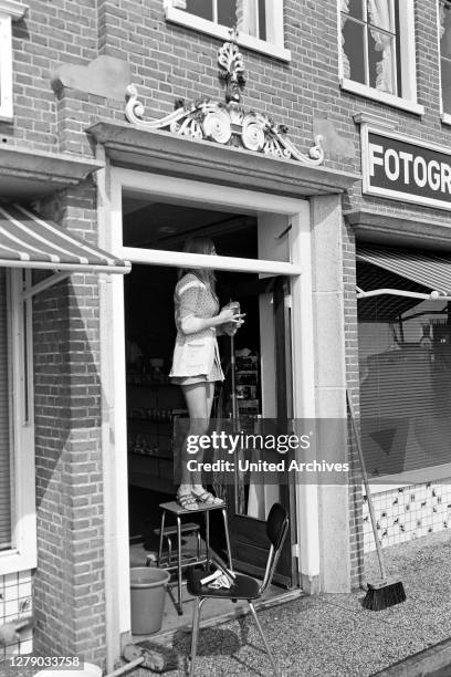 Young woman wearing a mini dress cleaning the windows of a photographic shop at Volendam, The Netherlands 1971.