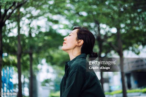 young asian businesswoman taking a break in urban park in the city. enjoying the gentle breeze with her eyes closed, resting in the middle of a work day - meditation stock pictures, royalty-free photos & images