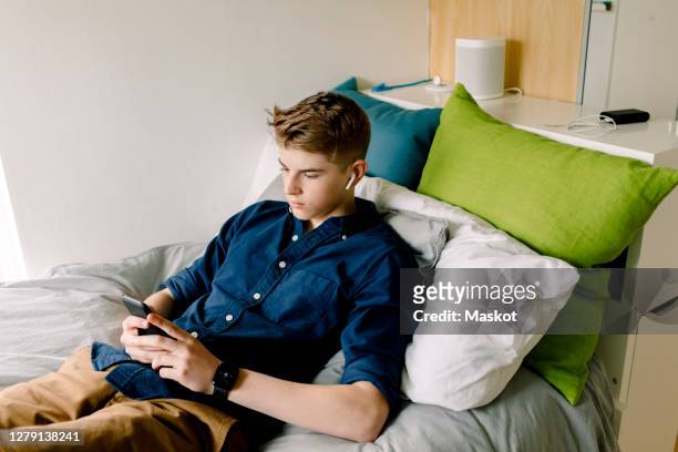teenage boy with in-ear  headphones using smart phone while sitting on bed at home - boy bedroom stock pictures, royalty-free photos & images