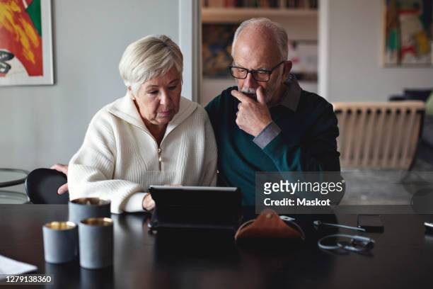 senior couple using digital tablet while sitting by dining table in living room - senior couple stock pictures, royalty-free photos & images