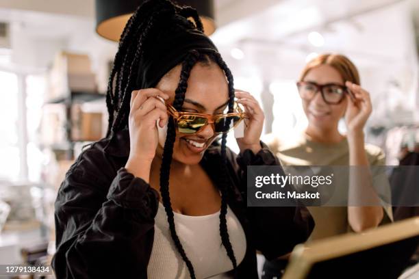 smiling female friends trying eyewear while talking at fashion store - sunglasses stock pictures, royalty-free photos & images