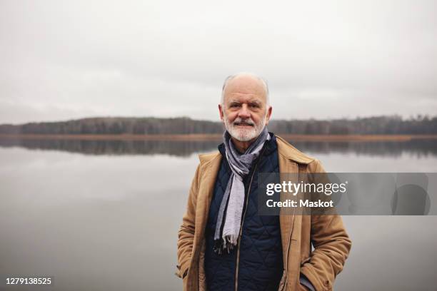 portrait of smiling senior man with hands in pockets standing against lake - jacket stock pictures, royalty-free photos & images