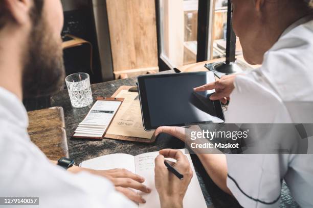 midsection of chef with digital tablet while coworker writing at table in restaurant - 50s woman writing at table imagens e fotografias de stock