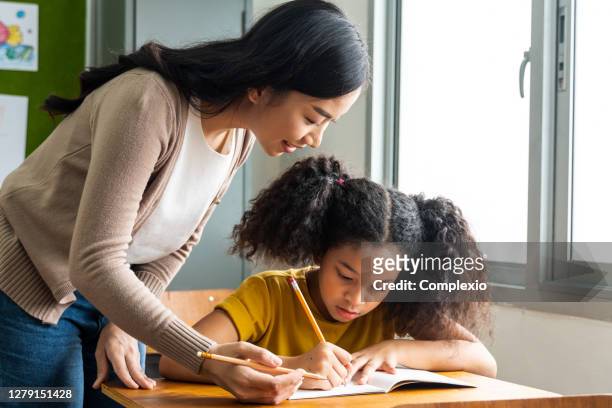 asian school teacher assisting female student in classroom. young woman working in school helping girl with her writing, education, support, care - teacher stock pictures, royalty-free photos & images