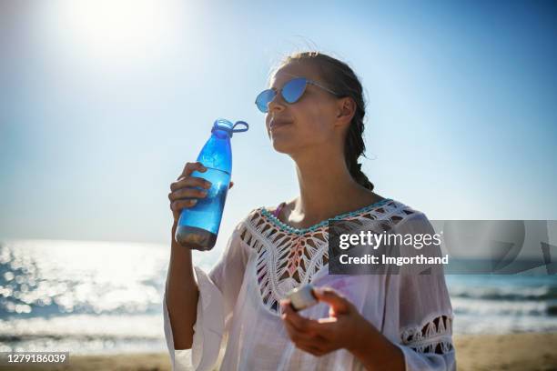 teenage girl on beach drinking from reusable water bottle. - blue sunglasses stock pictures, royalty-free photos & images