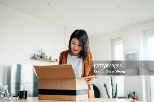 smiling young woman opening a delivery box in the living room - bulto fotografías e imágenes de stock