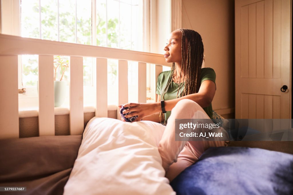 Young woman looking out of her bedroom window while drinking coffee