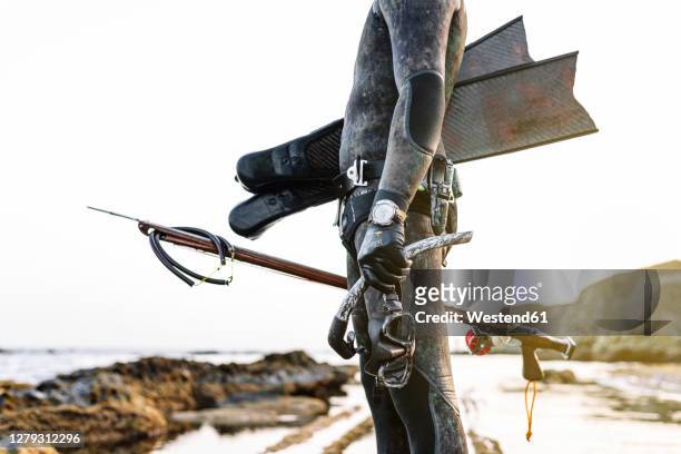 man holding scuba mask and harpoon while standing at beach - man spear fishing stock pictures, royalty-free photos & images