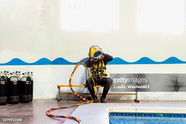 man wearing diving suit while sitting on bench against wall at poolside - immersione da palombaro foto e immagini stock