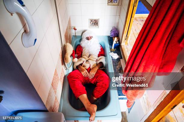 man wearing santa claus costume lying in bathtub at home - santa claus lying stock pictures, royalty-free photos & images