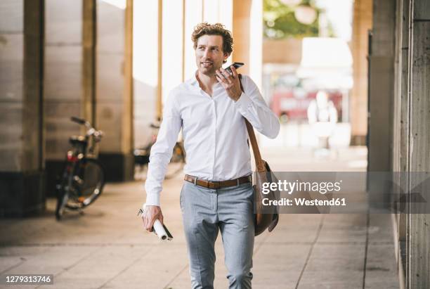 smiling man talking on phone while walking at building in city - business man looking at smart phone stock-fotos und bilder
