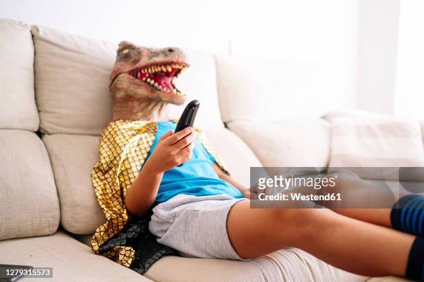 boy wearing dinosaur mask watching tv while relaxing on sofa at home - kind vor der pubertät stock-fotos und bilder