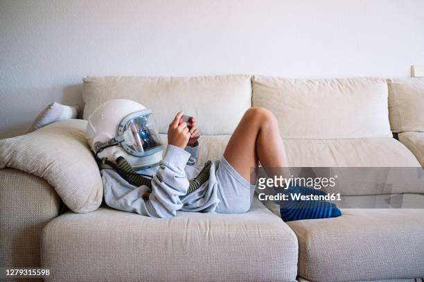 boy wearing space helmet playing video game while lying on sofa at home - astronaut kid stockfoto's en -beelden