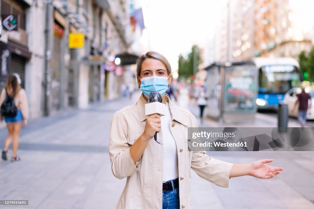 Female reporter wearing mask talking over microphone on street in city