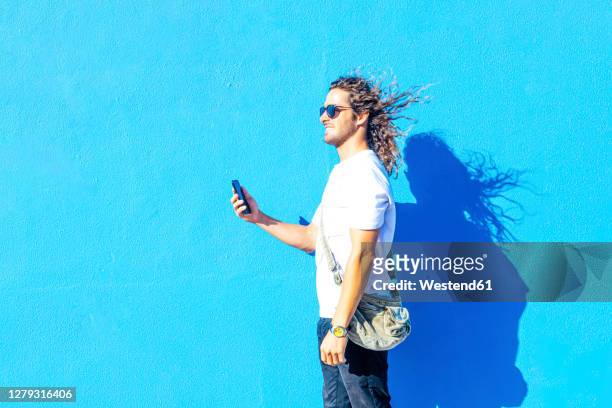 smiling young man wearing sunglasses using smart phone while standing by blue wall - hair wind stock pictures, royalty-free photos & images
