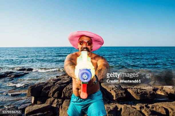 man wearing hat showing squirt gun while standing against sea - funny tourist stock pictures, royalty-free photos & images