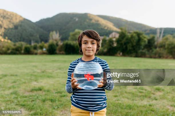 smiling boy holding fishbowl in hand while standing at back yard - pet goldfish stock pictures, royalty-free photos & images