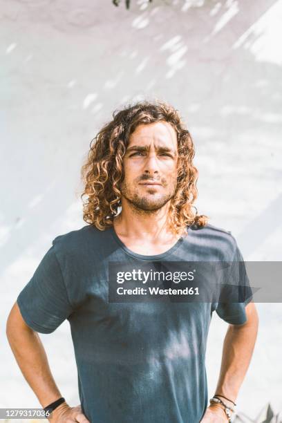 serious young man with hands on hip standing against white wall - long blonde hair men stock pictures, royalty-free photos & images