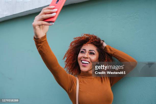 cheerful afro young woman taking selfie with smart phone while standing by wall - kinky stock-fotos und bilder