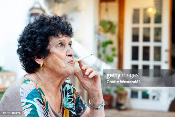 close-up of senior woman smoking cigarette while sitting outside house in yard - females smoking stock pictures, royalty-free photos & images