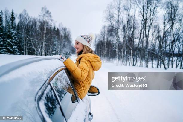 smiling mature woman sitting on car window against snow covered landscape - winter car window stock-fotos und bilder