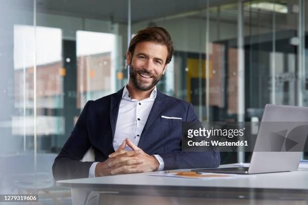 smiling businessman with hands clasped sitting by desk in office - business man at desk stock-fotos und bilder