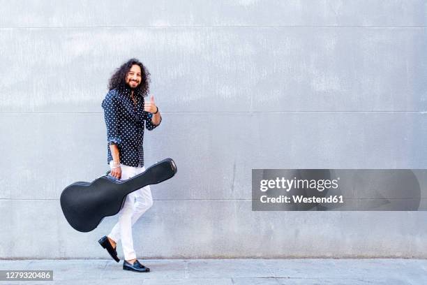 smiling man with guitar showing thumps up while walking against gray wall - guitar case fotografías e imágenes de stock