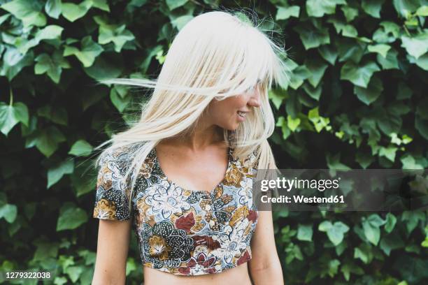 smiling young woman shaking hair while standing against plants in park - haare schütteln stock-fotos und bilder