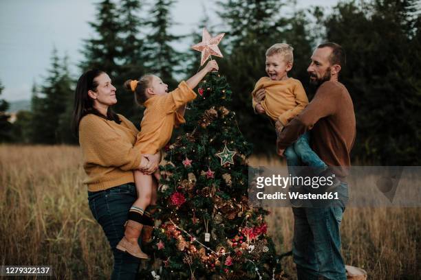 parents carrying happy children decorating christmas tree at countryside during sunset - kerstboom versieren stockfoto's en -beelden