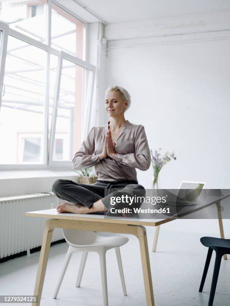 businesswoman meditating while sitting on desk in loft office - schneidersitz stock-fotos und bilder
