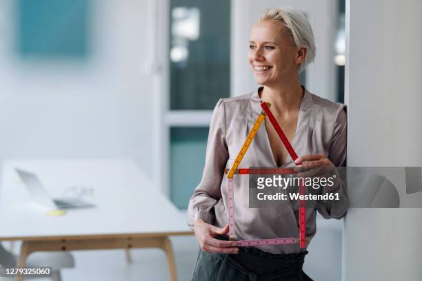 cheerful female architect holding model home while standing by wall in office - architekturmodell stock-fotos und bilder